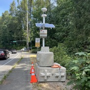 An electrical device with a solar panel on top of a concrete block, next to a sidewalk and street