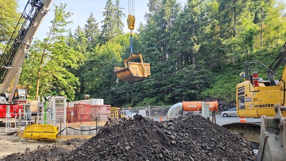 A crane lifting dirt over an active construction site with trees in the background