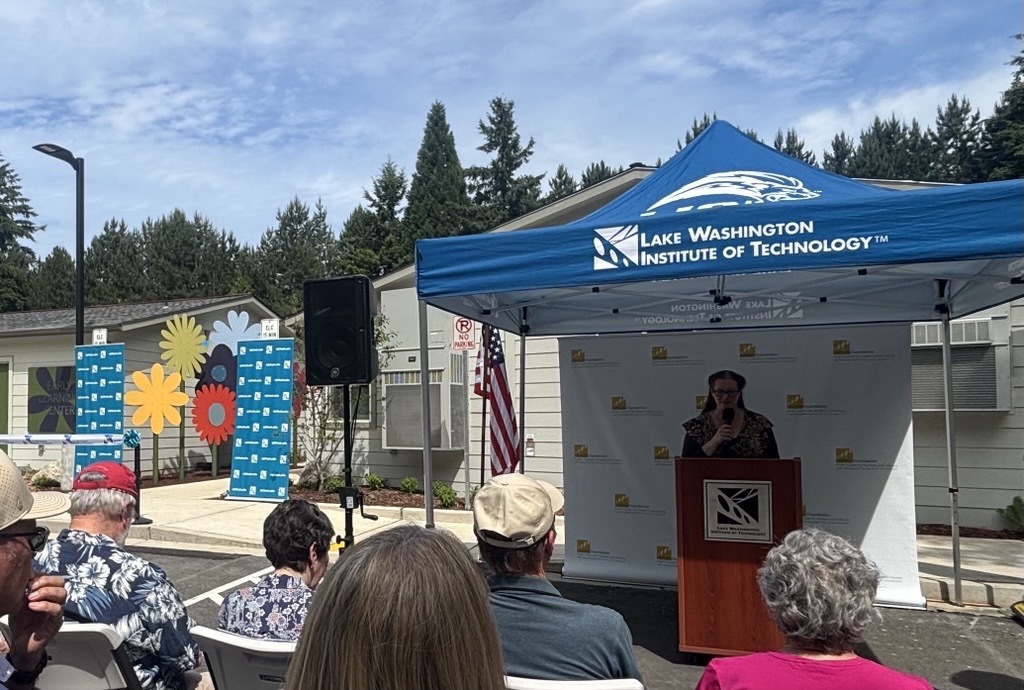 A speaker stands under a blue canopy speaking to a crowd with low beige buildings, trees, and a blue sky in the background