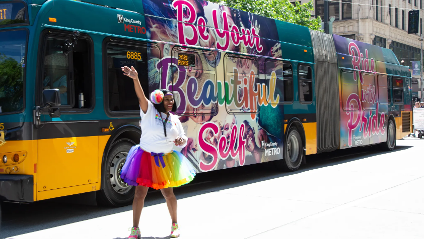 image of the pride bus wrapped with pride images and a smiling metro employee out in front of it in a parade 