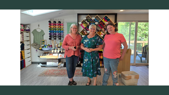 Three women stand, smiling, in front of a white wall lined with shelves of colored yarn.