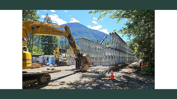 An excavator sits in front of the metal framework of a bridge.