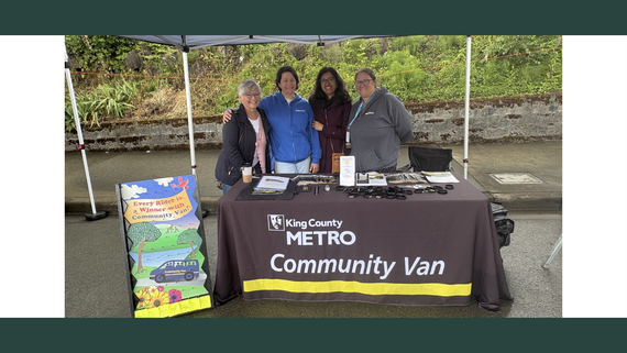 Four people stand behind a table covered in a black cloth with white text that says King County Metro Community Van.