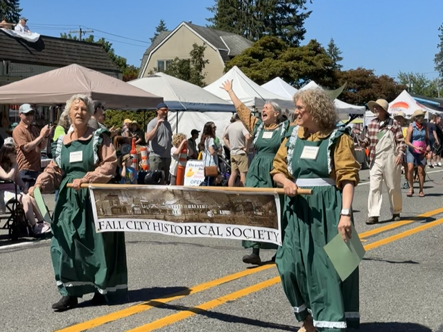 Three women in old fashioned green and yellow dresses march down a street with a banner reading Fall City Historical Society.