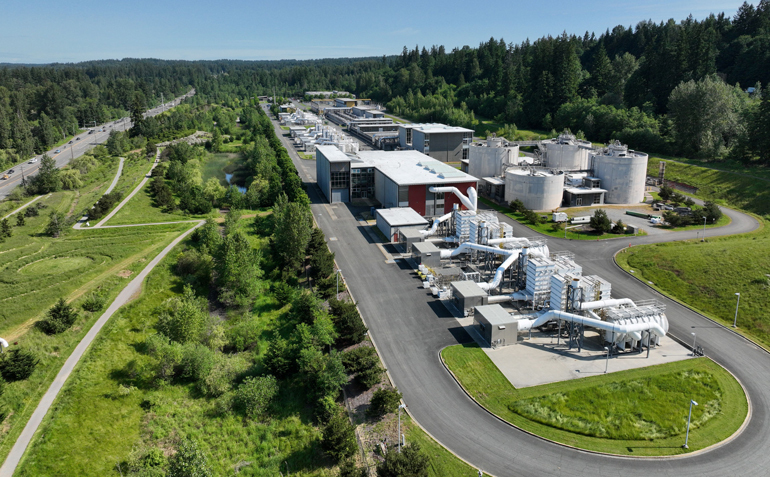 White buildings and machinery surrounded by a gray road and green grass and trees against a blue sky
