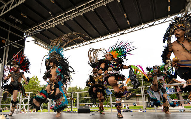 Six dancers in brown costumes and colorful feather headdresses on a stage under a black ceiling against a gray sky