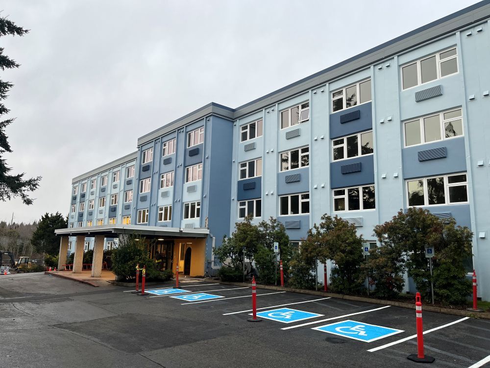 A four story blue building with small trees in front of it and a parking lot with blue and white painted ADA parking spots