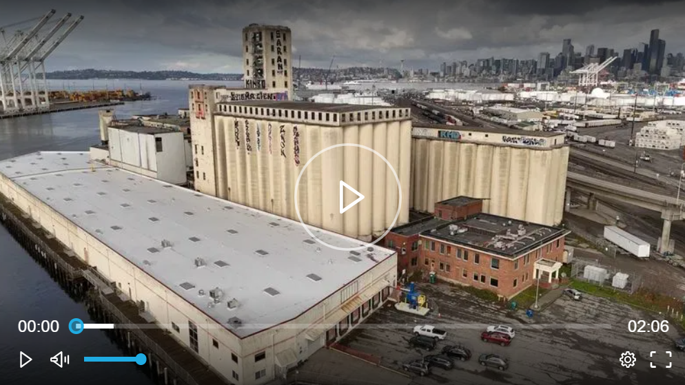 Aerial shot of industrial buildings and grain silos in front of a waterfront and a city skyline