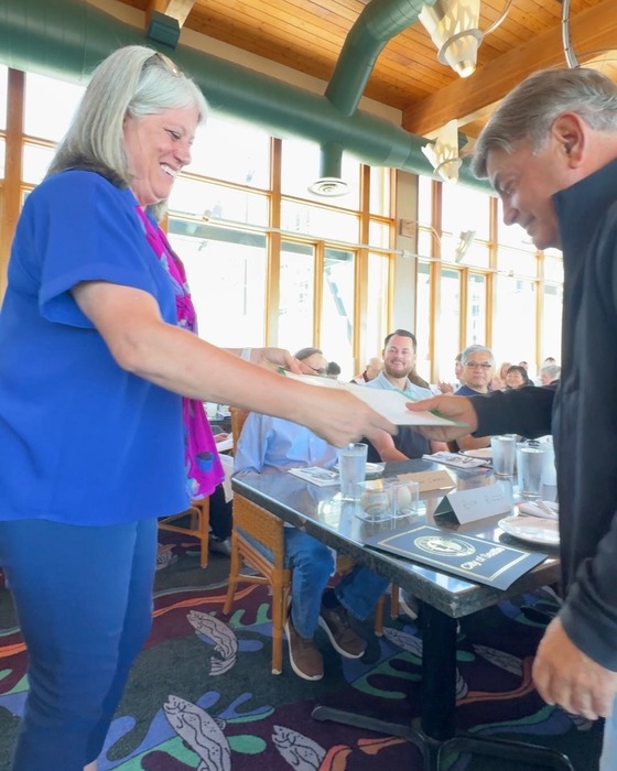 A woman in blue hands a document to a man in a black jacket with people sitting at a table behind them