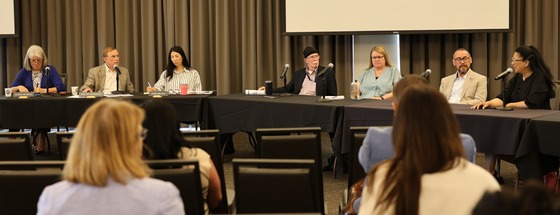 A panel of 7 individuals seated at a table with microphones, speaking to an audience at the Museum of Flight. 