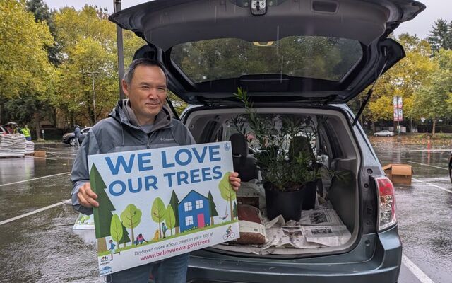 A man stands in a parking lot with a sign saying "We love our trees" in front of an open car trunk with small trees in it