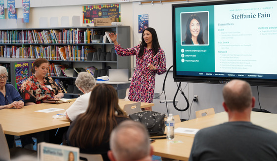 Councilmember Fain speaking in a library in front of people and a screen behind her with her photo and bio