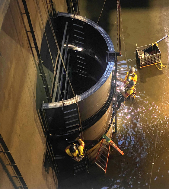 Two workers wearing protective gear climbing a ladder attached to a tall, curved metal form suspended by chains.