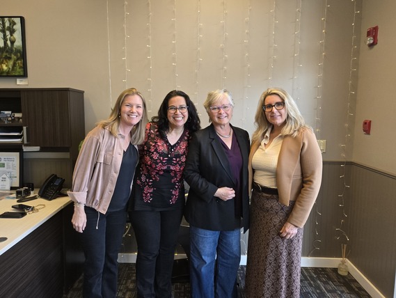Four women stand, smiling, in front of a beige wall with white string lights hanging down from the ceiling.