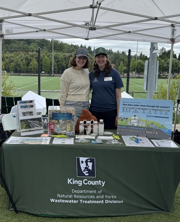  Two employees stand behind a table with a King County logo and information about Brightwater and wastewater treatment.