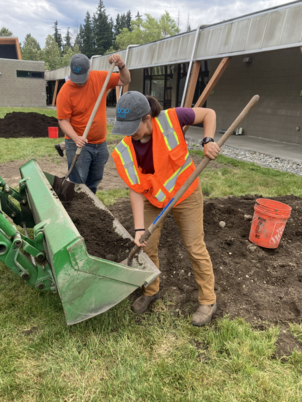 Two people in orange vests and Loop hats use shovels to transfer piles of Loop compost and dirt into a green tractor.