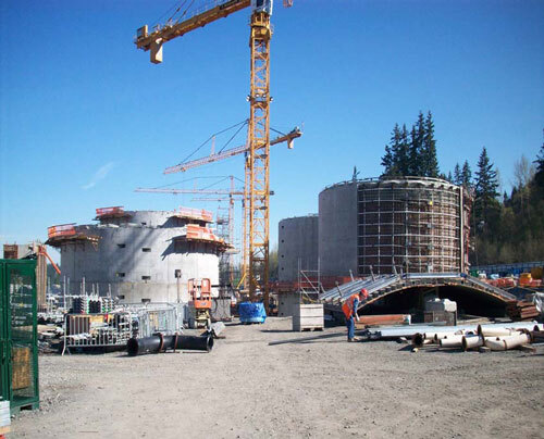 Large yellow cranes tower over large, oval, concrete structures. In the foreground, pipes are visible on the dirt-covered ground. 