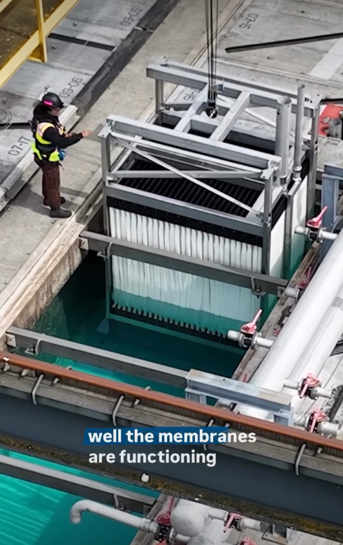 Aerial view of a utility worker using a crane to maneuver a new membrane cassette into a tank of clear blue water.