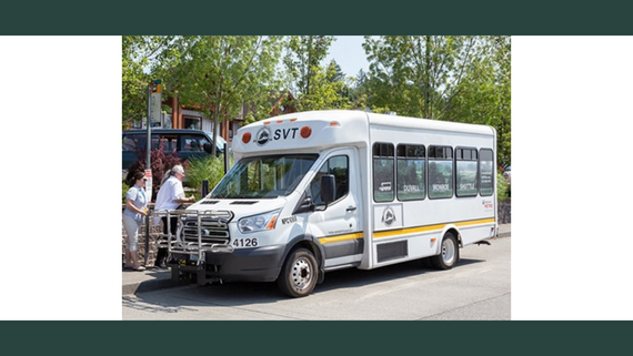 A small white bus with black letters painted on top reading SVT picking up people from a bus stop.
