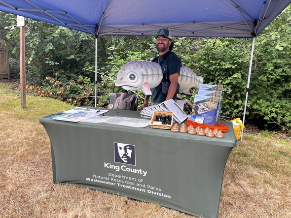 Man behind a information table wearing a salmon costume around his torso.