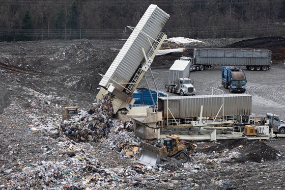 A large machine tips its a metal container up to unload trash onto the ground at a landfill as trucks carrying more containers approach..