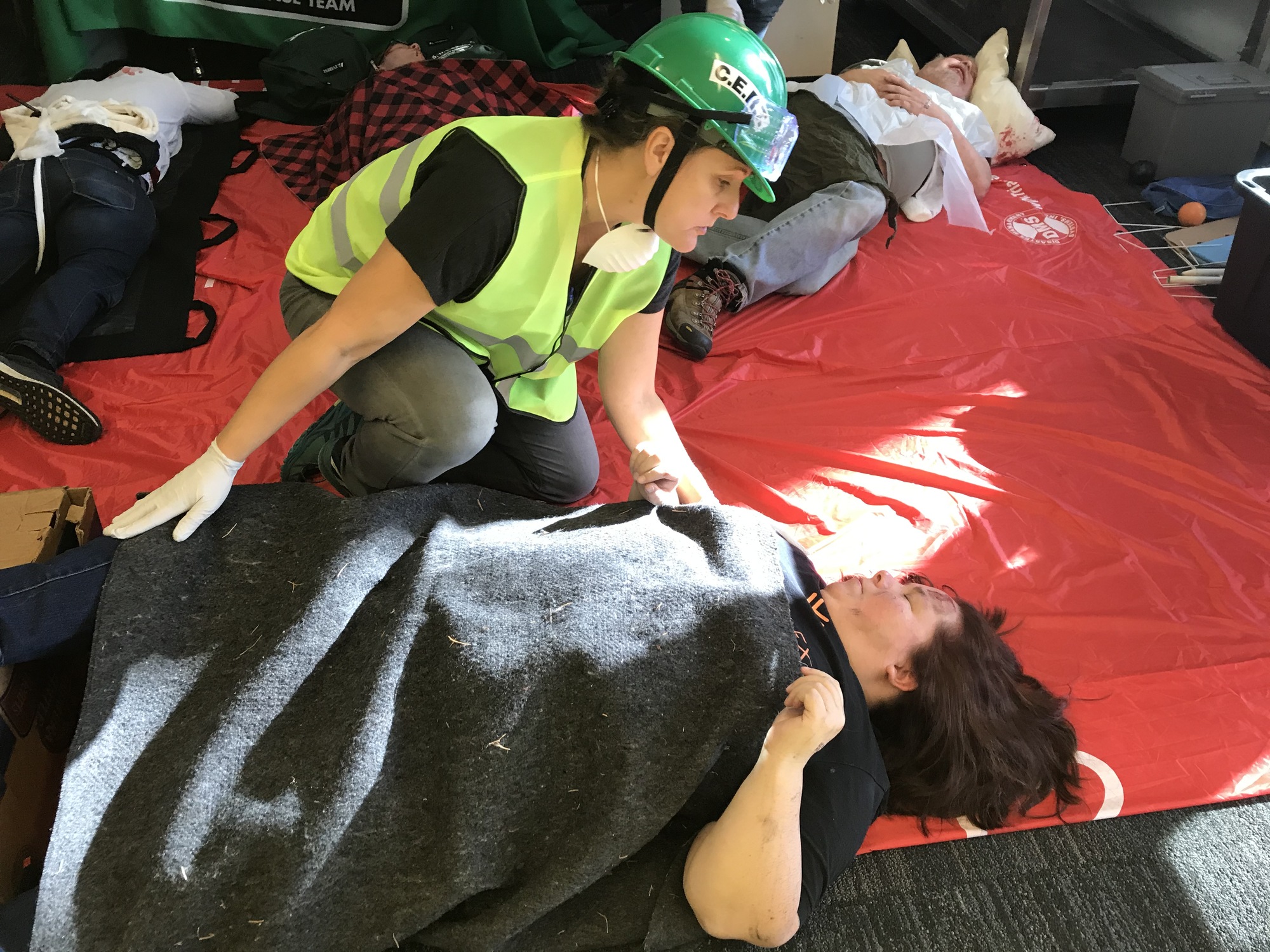 A person in a green CERT vest and helmet reaches out to a volunteer earthquake survivor lying on a red tarp during a CERT exercise.