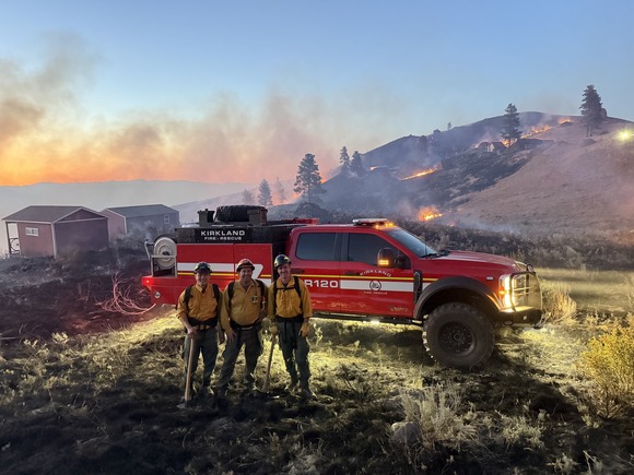 Image shows Kirkland firefighters in front of a firetruck with wildfire in the distance