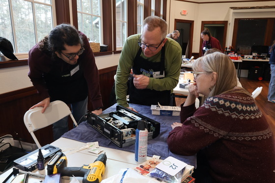 Two volunteer fixers helping to repair a computer tower while a participant watches.