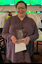 Vicky Schiantarelli, a brown haired woman wearing glasses in a floral patterned button up shirt holding an award that says BCCAP