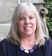 Jill Diehl, photo of smiling womani n front of of gray building wall with greenery in the background.