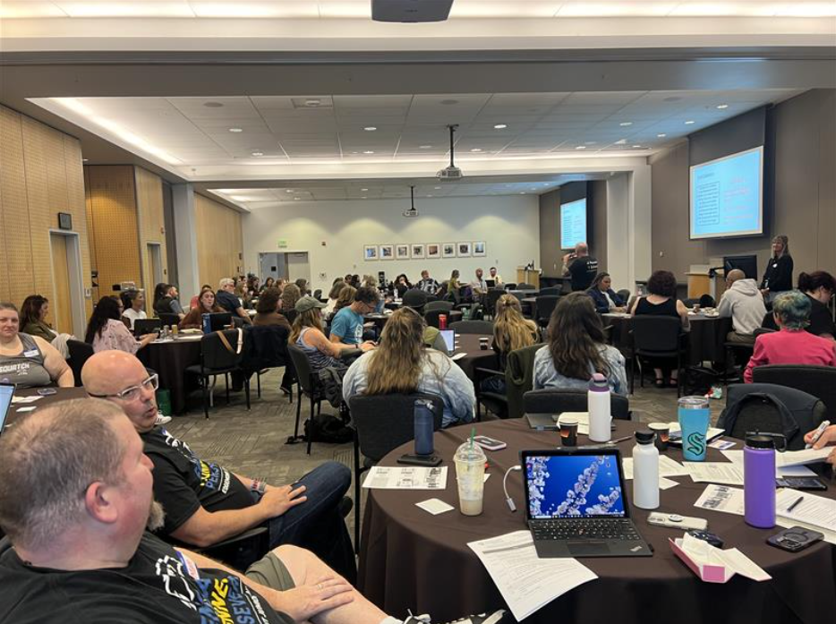 Attendees at the mastery-based learning Spring 2026 gathering sit at tables and listen to a presentation