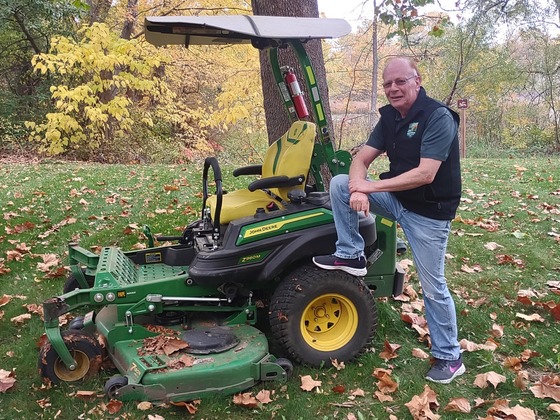 A smiling male park aide stands next to a tractor lawn mower with golden fall trees behind him.