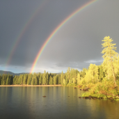 A double rainbow above a lake in front of a dark cloud; sun reflects off the trees and water.