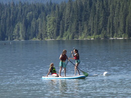 Three children play on standup paddleboards in a lake, wearing life jackets.