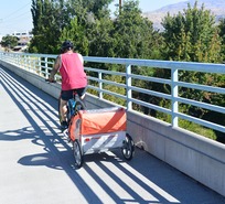 A tattooed bicyclist tows an orange child trailer on his bike, over a bridge with long shadows casting patterns