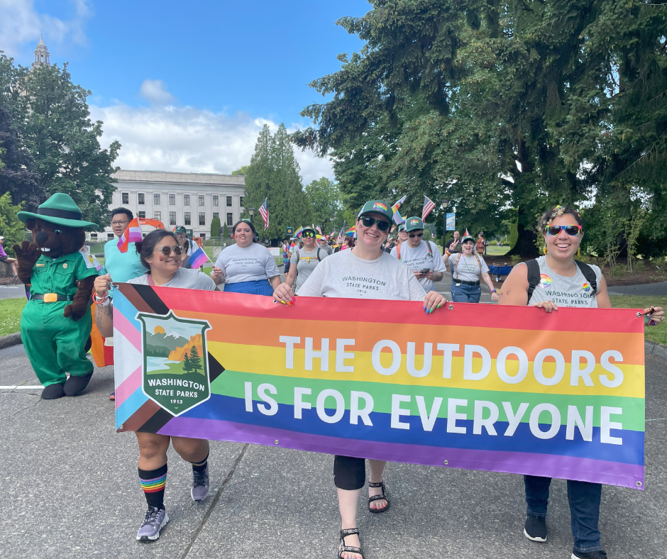 Parks staff walking at the Olympia Pride Parade holding a poster that says The Outdoors is for Everyone