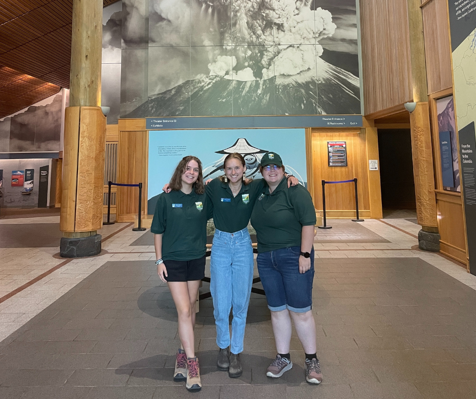 Three past interns standing in front of the erupting Mount St. Helens photo at the visitor center