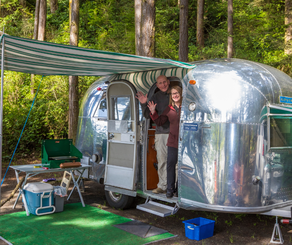 Two people poking their heads out and waving from their airstream trailer at Deception Pass