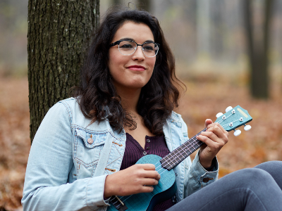 young woman playng ukulele outside, leaning against a tree in the forest