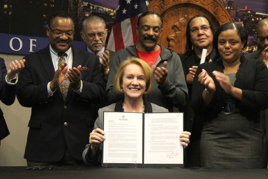 Mayor Durkan holds up the signed WMBE Executive Order with several small business owners clapping in the background
