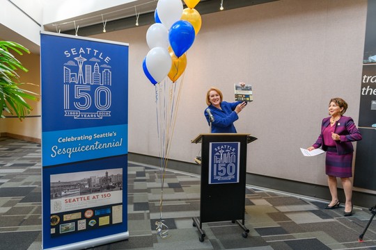 Mayor Jenny Durkan holds the Seattle at 150 book above her head in celebration with City Clerk Monica Martinez Simmons looking on