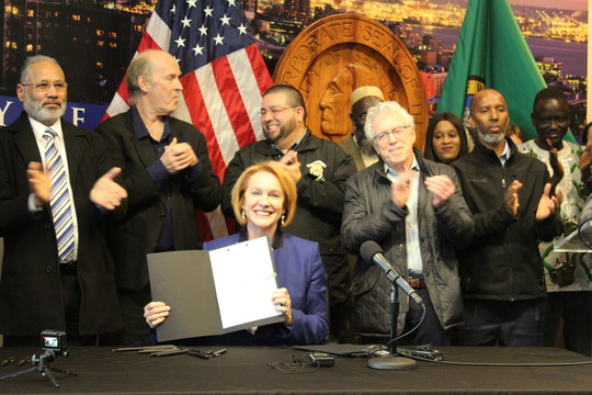 Mayor Durkan holds signed Fare Share legislation up while labor advocates and drivers applaud in the background. 