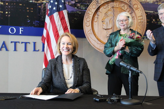 Mayor Durkan smiles after signing the 2020 budget. CM Sally Bagshaw holds a bouquet of flowers in the background.