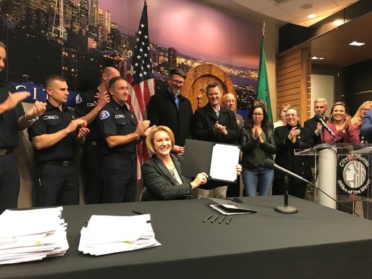 Mayor Durkan holds a signed contract up and smiles at the camera, surrounded by smiling City employees