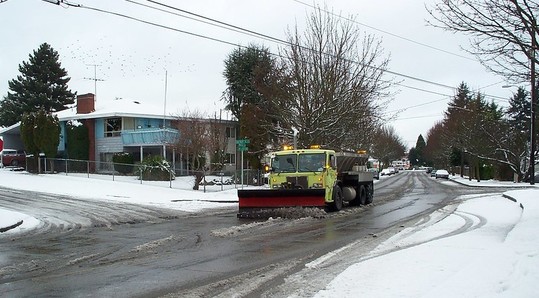 A snow plow scoops up snow on a wide street covered in slush.