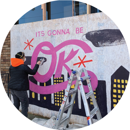 artist painting mural on plywood covering storefront