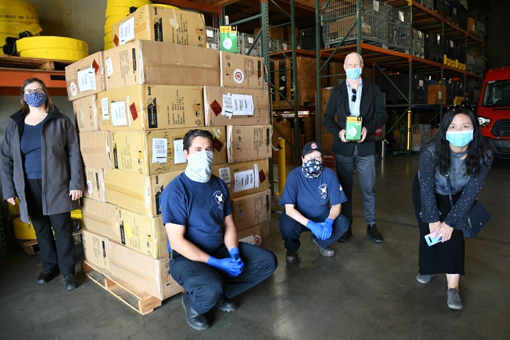 Photo of individuals wearing PPE masks in front of a large stack of donated PPE