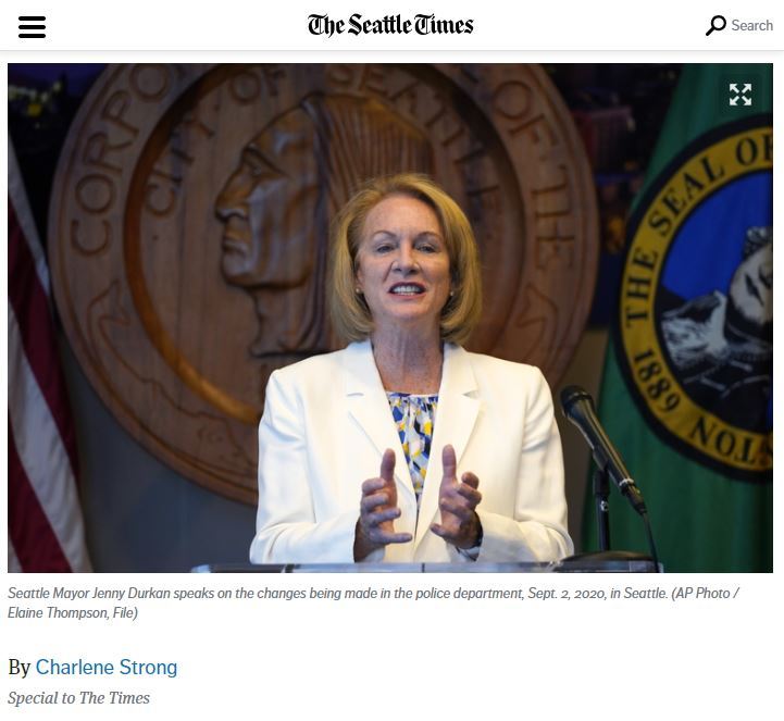 Photo of Mayor Durkan in a white blazer in front of the City of Seattle seal and flags