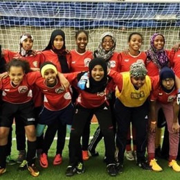 A group of East African teenage girls with arms around each others, smiling and standing in front of a soccer goal.