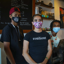 Three people wearing face masks stand behind the counter of a coffee stand with a chalkboard menu on the wall behind them.